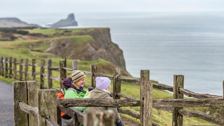 Two people sat on a bench wearing coats and hats. There is a wooden fence between them and they are overlooking the see with the hills in the background